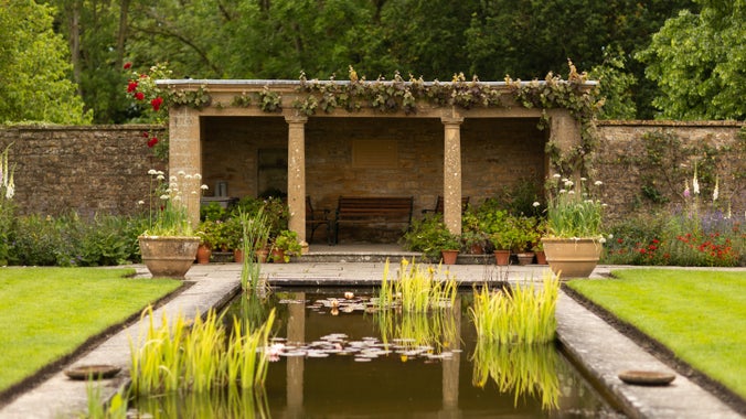 A rectangular pool with reeds in front of a stone pergola with planted pots and a bench.